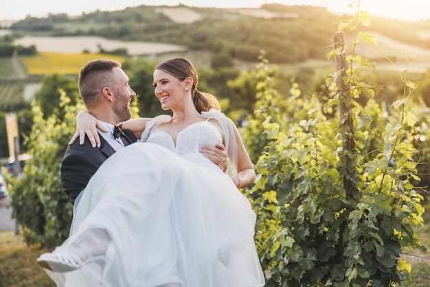 Foto sposo allegro che trasporta e gira la sua sposa mentre si gode in vigna il giorno delle nozze.