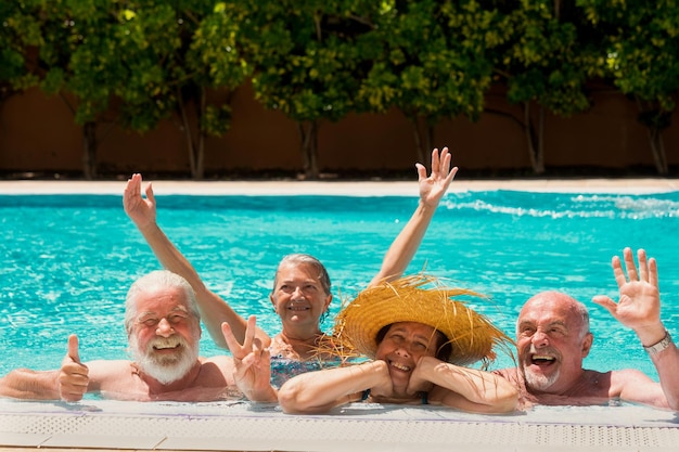 Cheerful group of four senior people having fun in the outdoor swimming pool Caucasian elderly men and women smile relaxed on vacation under the bright sun