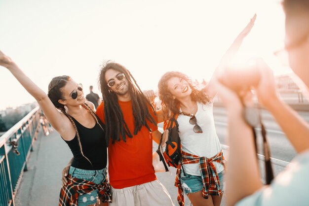 Cheerful group of multicultural tourists posing on the bridge while their friend taking photo of them. Summer holiday concept.