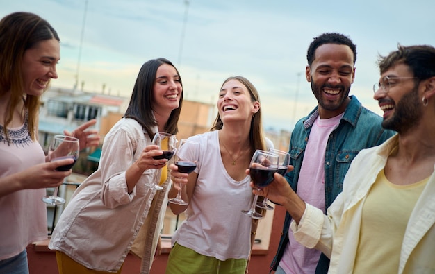 Cheerful group of multiracial young friends laughing holding wine glass at summer rooftop party