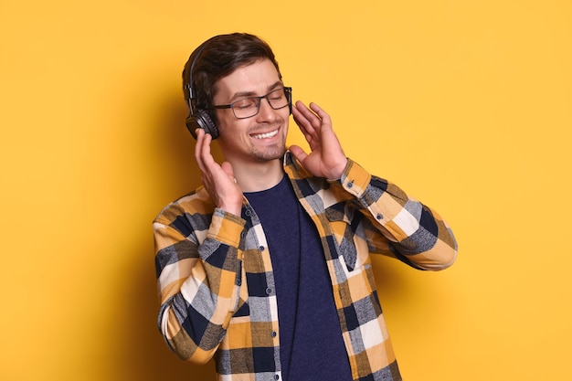 Cheerful guy listening favorite music with headphones against yellow studio wall