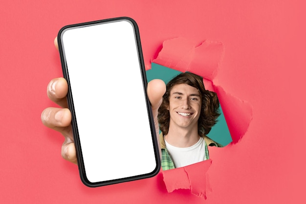 Cheerful guy showing white empty smartphone screen through torn paper