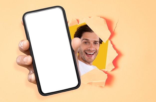 Cheerful guy showing white empty smartphone screen through torn paper