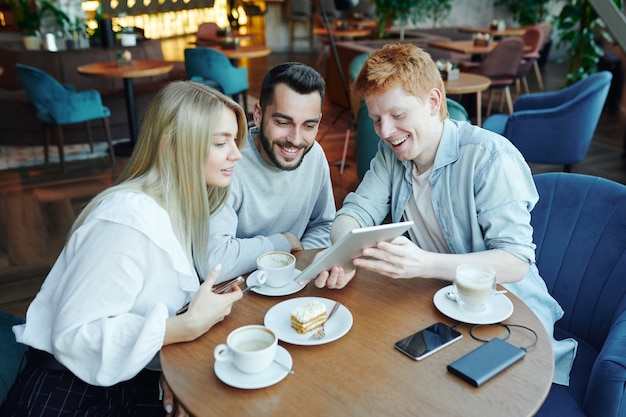 Ragazzo allegro con il touchpad che mostra ai suoi amici cose curiose mentre è seduto a tavola nella caffetteria con una tazza di caffè dopo le lezioni