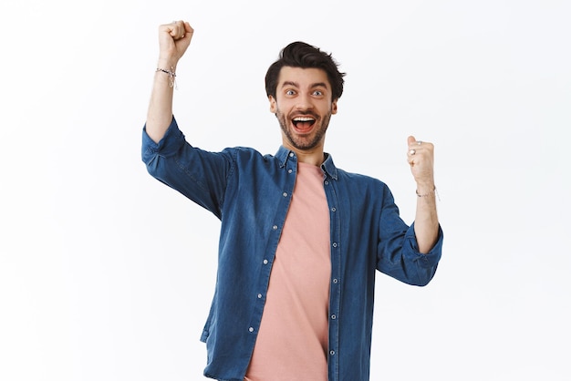 Cheerful handsome bearded man raising arms up in hooray congratulations gesture fist pump as triumphing winning watching intense football game smiling relieved and happy stand white background