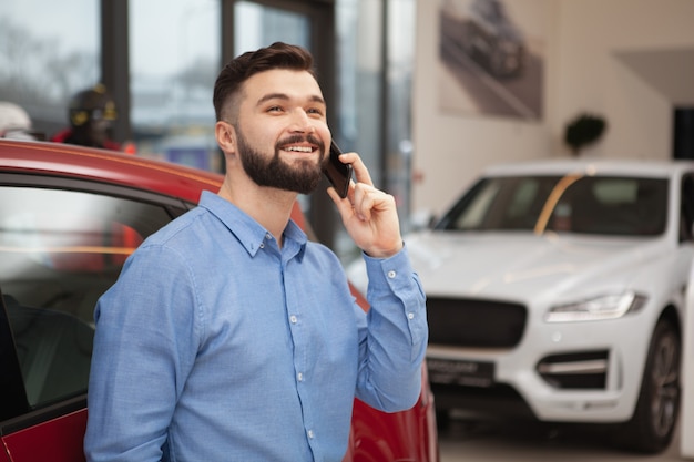 Cheerful handsome bearded man smiling cheerfully, looking away while talking on the phone at car dealership, copy space