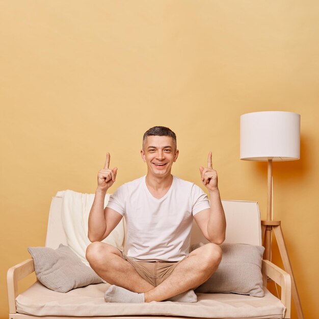 Cheerful handsome brunette adult man wearing casual clothing sitting on sofa against beige wall pointing upwards with index fingers advertisement area