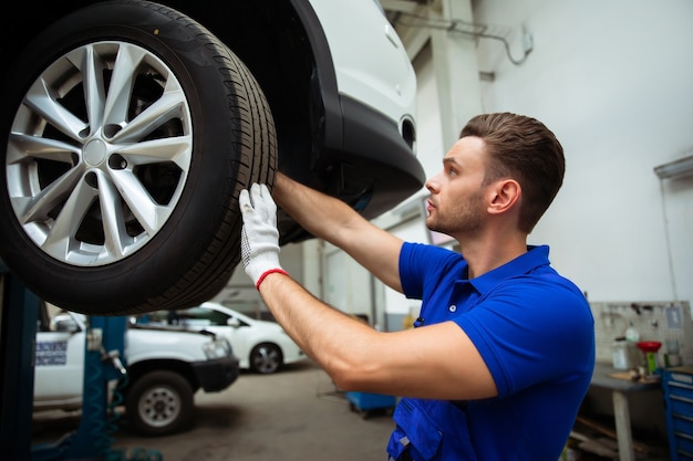 Cheerful, handsome, and confident car repair specialist in overalls repairs and replaces old parts with new ones in a car on a lift in service