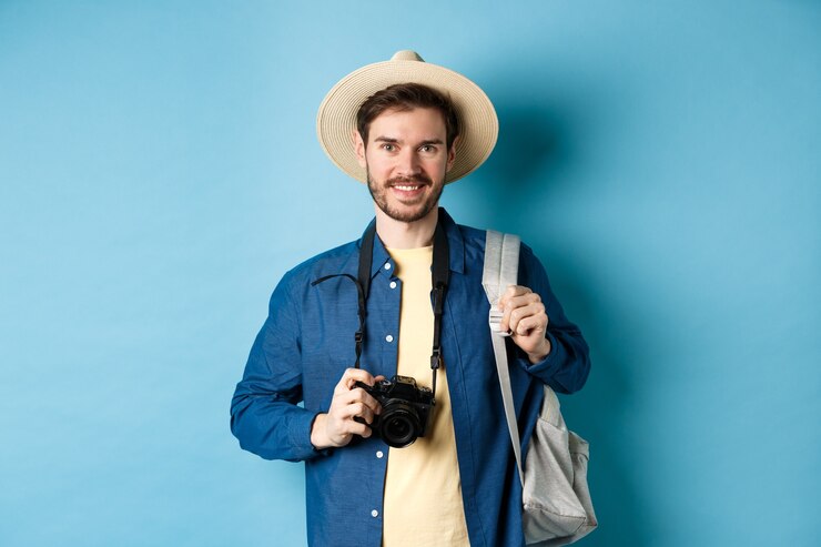 Photo cheerful handsome guy going on vacation, wearing summer hat and holding backpack with camera for photos, smiling excited of holiday, standing on blue background.