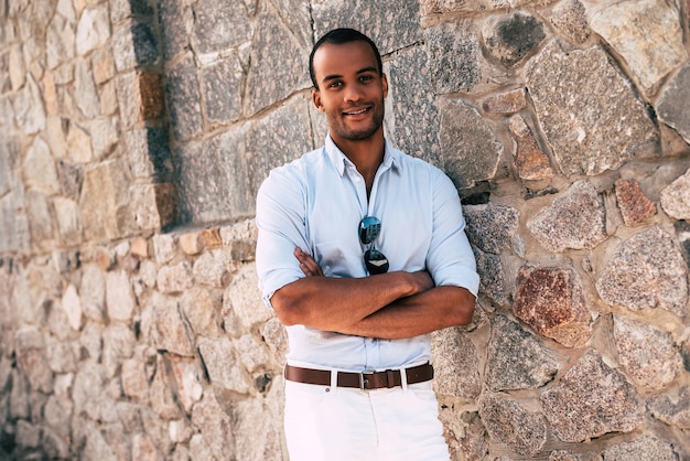 Cheerful and handsome. Handsome young African man in smart casual clothes keeping arms crossed and looking at camera with smile while standing against the stoned wall outdoors