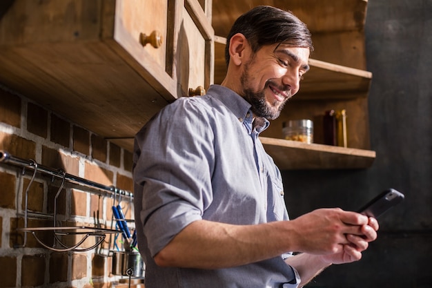 Cheerful handsome man resting in the kitchen while using his smartphone