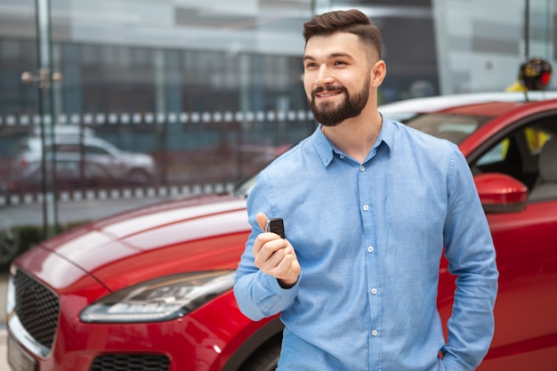 Cheerful handsome man smiling, looking away, holding keys to his new automobile at car dealership