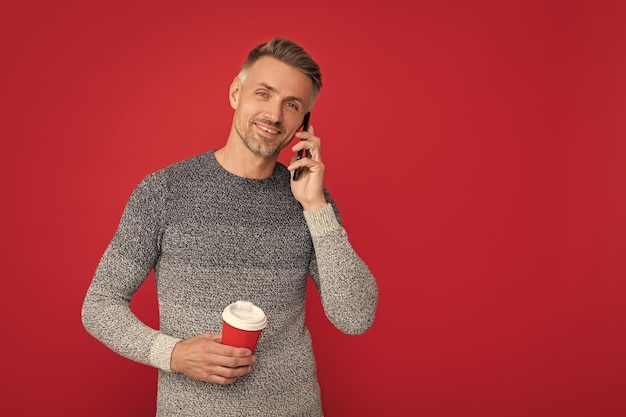 쾌활한 잘 생긴 남자는 스웨터를 입고 전화와 커피 컵 대화를 들고