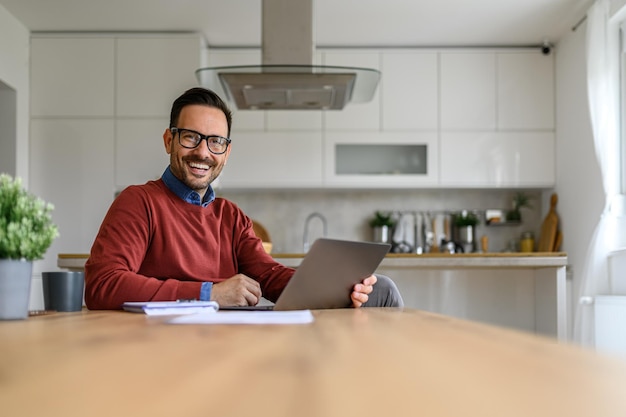 Cheerful handsome man with laptop and note pad on desk working from home office smiling at camera