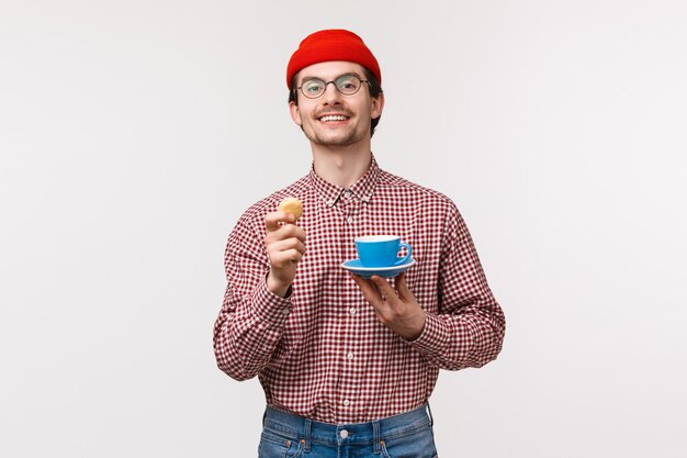 Cheerful handsome and relaxed young caucasian bearded man enjoying his tea with biscuits, holding cookie and cup as drinking coffee during lunch break, smiling pleased, 