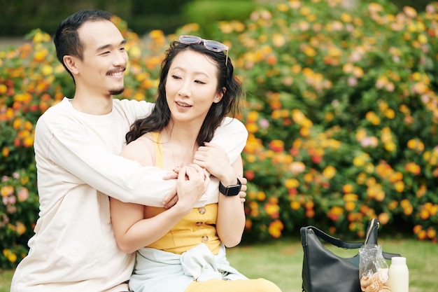 Cheerful handsome young Chinese man in love hugging his girlfriend when they are resting on bench in park