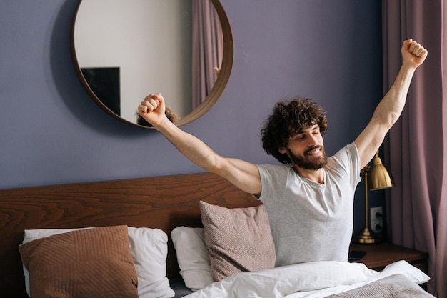 Фото Веселый красивый молодой человек просыпается и протягивает руки утром, сидя на кровати счастливый бородатый мужчина видит приятный сон