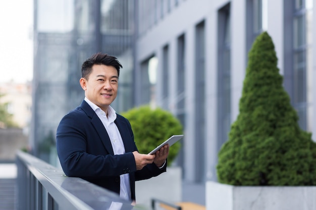 Cheerful and happy asian boss reads news from tablet near modern office male businessman in business suit smiles from success