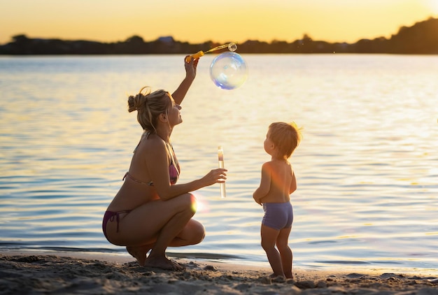 Cheerful happy beautiful mother in modern bikini swimsuit plays with her little funny baby inflating bright multicolored soap bubbles for him, while enjoying sunny summer holidays