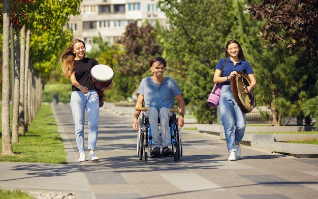 Cheerful. Happy caucasian handicapped man on a wheelchair spending time with friends playing live instrumental music outdoors. Concept of social life, friendship, possibilities, inclusion, diversity.