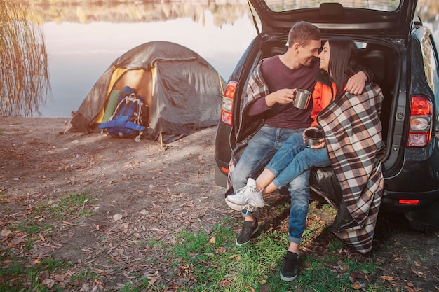 Cheerful and happy couple sitting together close to each other. They look and smile. Young woman is covered partly with blanket. Couple sits in trunk at water.