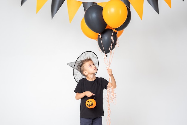 A cheerful happy Halloween boy in a wizard's hat holding a pumpkinshaped bucket and black and orange balloons