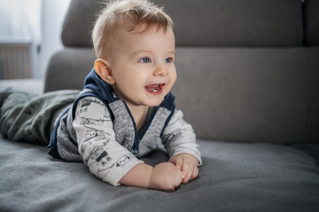 Cheerful happy little baby boy smiling and lying on stomach on couch in living room.