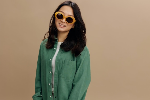 Cheerful happy lovely korean young woman in khaki green shirt\
stylish eyewear smiles at camera posing isolated on over beige\
pastel studio background cool fashion offer sunglasses ad\
concept