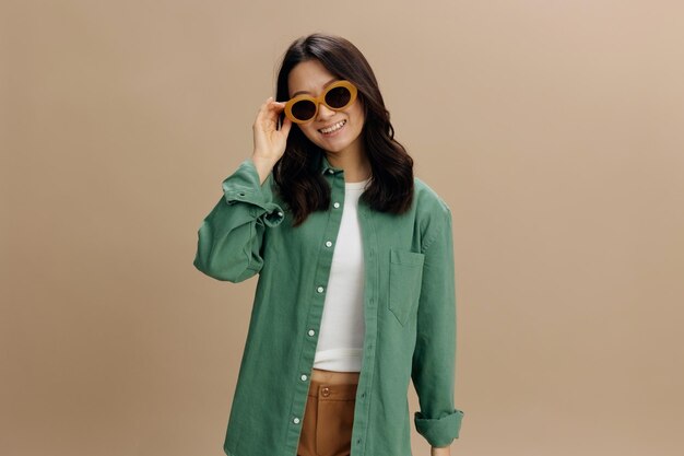 Cheerful happy lovely korean young woman in khaki green shirt\
stylish eyewear smiles at camera posing isolated on over beige\
pastel studio background cool fashion offer sunglasses ad\
concept