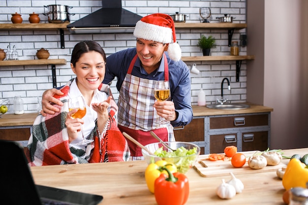 Cheerful happy man and woman celebrating Christmas or new year. Sitting together in room and smiling. Looking at laptop. Man wear hat. Hold wine glasses in hands.