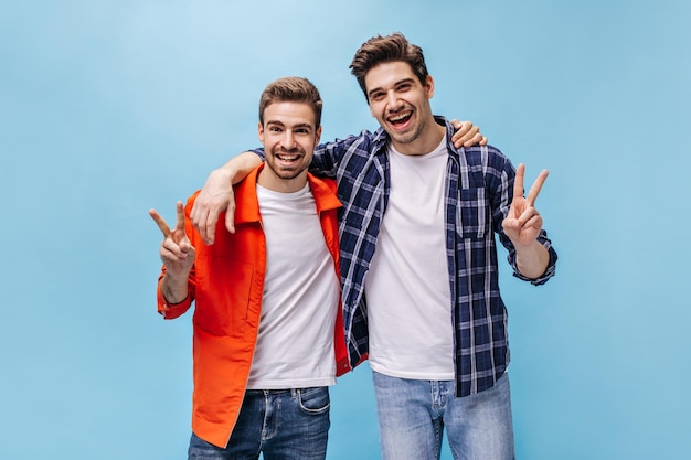 Cheerful happy men show peace signs on blue background Man in orange jacket and his friend in checkered shirt smile on isolated