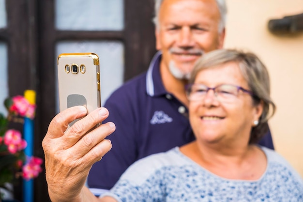 Cheerful happy old mature adult man and woman taking selfie with the phone at home smiles and happiness using phone to communicate with parents and son or daughters video conference and live call