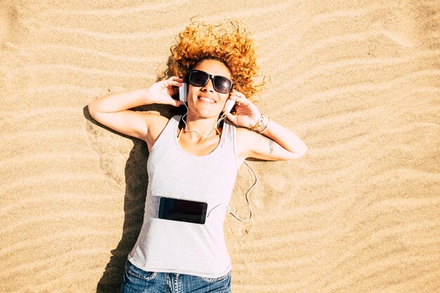 Cheerful happy woman lay down at the beach listinging music with phone and headphones enjoying summer travel holiday vacation alone in top view - happy female people portrait