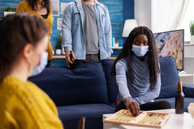 Cheerful happy woman playing backgammon with multiethnic friends wearing face mask as prevention for covid 19 spread, during global pandemic sitting on couch drinking beer and eating popcorn.