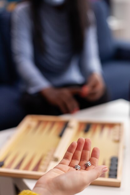 Cheerful happy woman using dice playing backgammon with multiethnic friends wearing face mask as prevention for covid 19 spread, during global pandemic sitting on couch drinking beer and eating popcor