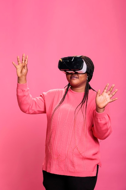 Cheerful happy woman wearing virtual reality headset playing video games enjoying 3d entertainment during lesiure time. Happy cheerful person with vr headset while standing over pink background