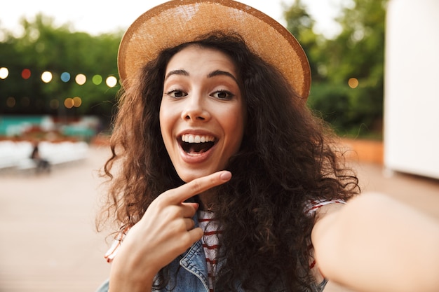 Cheerful happy woman with curly brown hair, smiling and pointing finger aside while taking selfie photo outdoor