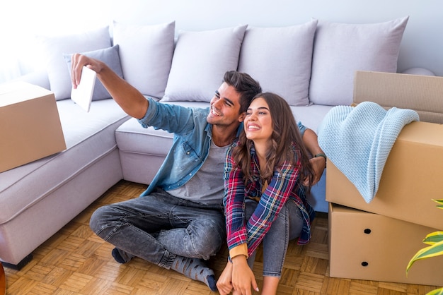 Cheerful and happy young couple making a selfie in new home with moving cardboard box duri