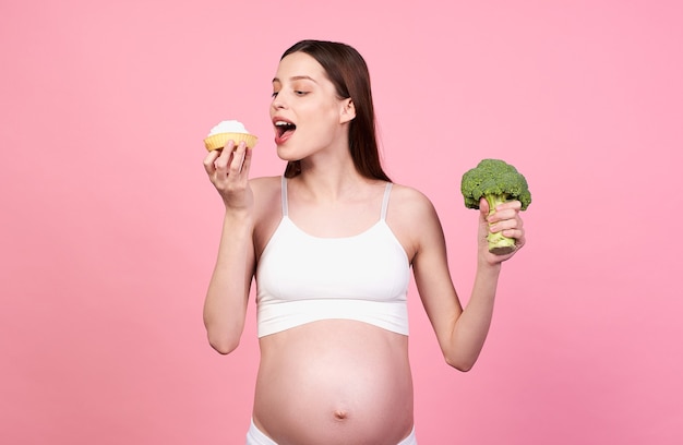 Cheerful happy young pregnant girl, in a white top and white leggings, greedily bites a beautiful cake, does not follow a diet for pregnant women. The concept of pregnancy and nutrition, diet.