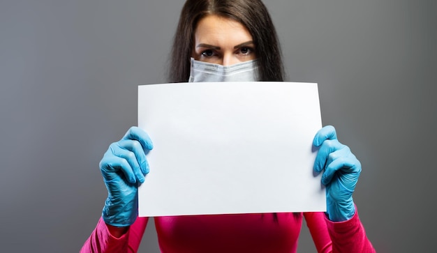 cheerful and healthy young woman wearing medical mask and gloves holding a empty sheet of paper to prevent others from corona COVID-19 and SARS cov 2 infection, copy space for individual text
