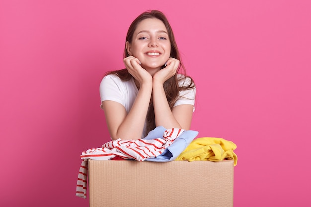 Cheerful helpful volunteer standing isolated over pink, holding box with clothes for secondary use, keeps hands under chin, looks at camera with pleased facial expression.