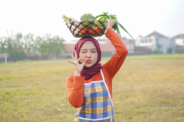 陽気なヒジャブの女性が野菜かごを頭の上に持ち上げる