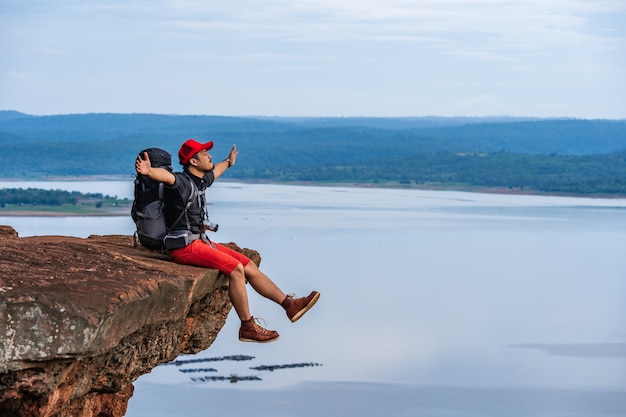 Cheerful hiker man sitting and gesture raised arms on the edge of cliff, on a top of the rock mountain