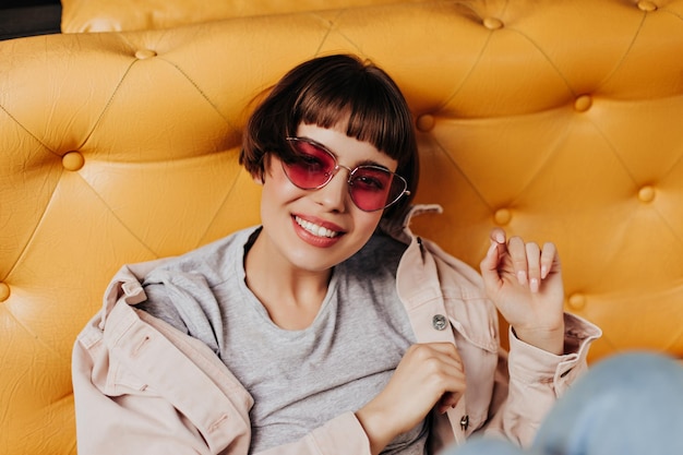 Cheerful hipster girl sitting on yellow couch inside Shorthaired woman in pink glasses grey tshirt and denim jacket smiling in cafe