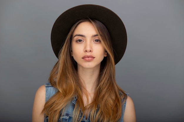 Cheerful hipster girl in stylish hat and denim vest posing at grey copy space wall for your content