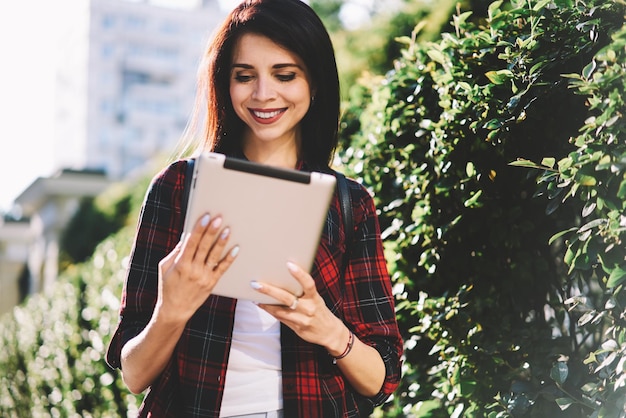 Ragazza allegra hipster che digita messaggi di testo e cerca informazioni sul percorso turistico tramite tablet