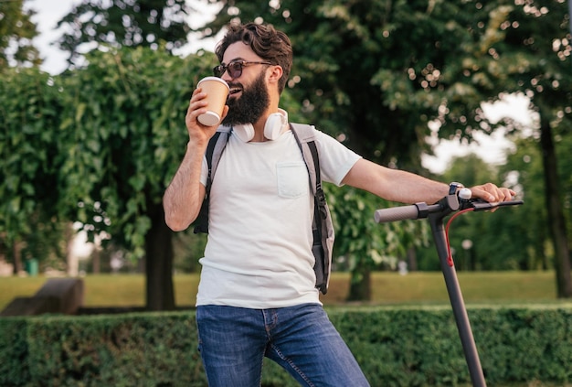 Cheerful hipster with scooter drinking hot beverage in park