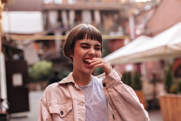 Cheerful hipster with short hair in light denim jacket laughing outdoors Humored brunette girl with beige outfit has fun in city