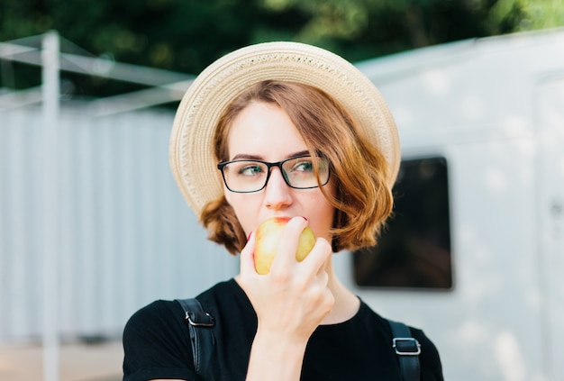 Foto donna allegra hipster in occhiali e cappello che mangia mela all'aperto.