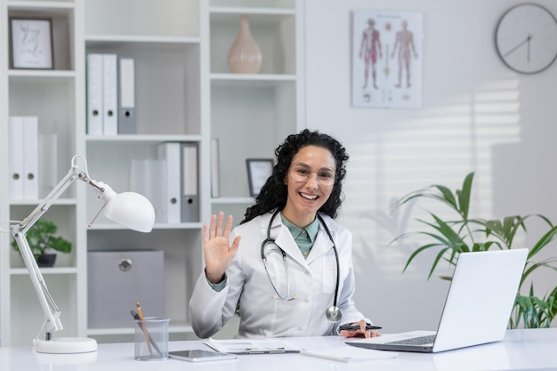 Веселая латиноамериканская женщина-доктор махает рукой в приветствии в своем офисе, представляя доступность